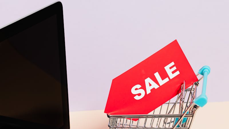 Miniature shopping cart with a sale tag next to a laptop, representing the everyday online purchases that drain your wallet